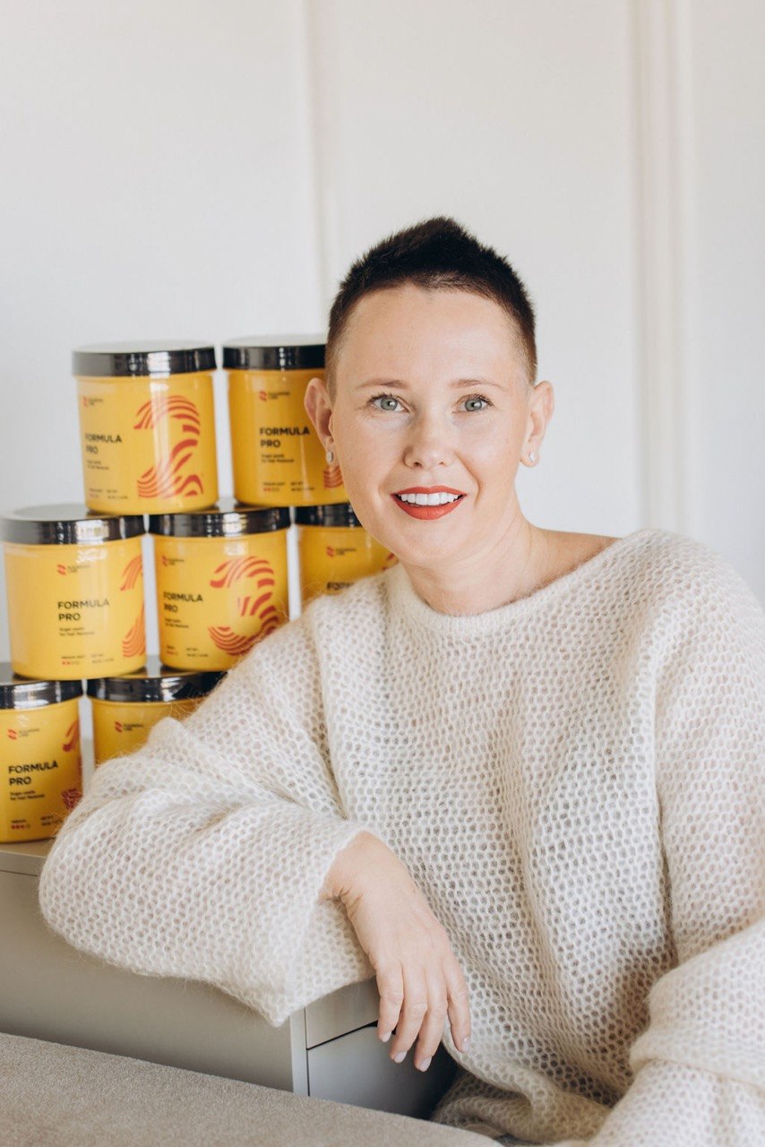 Woman with short dark hair and red lipstick smiling, sitting in front of stacked yellow containers, wearing a white sweater.