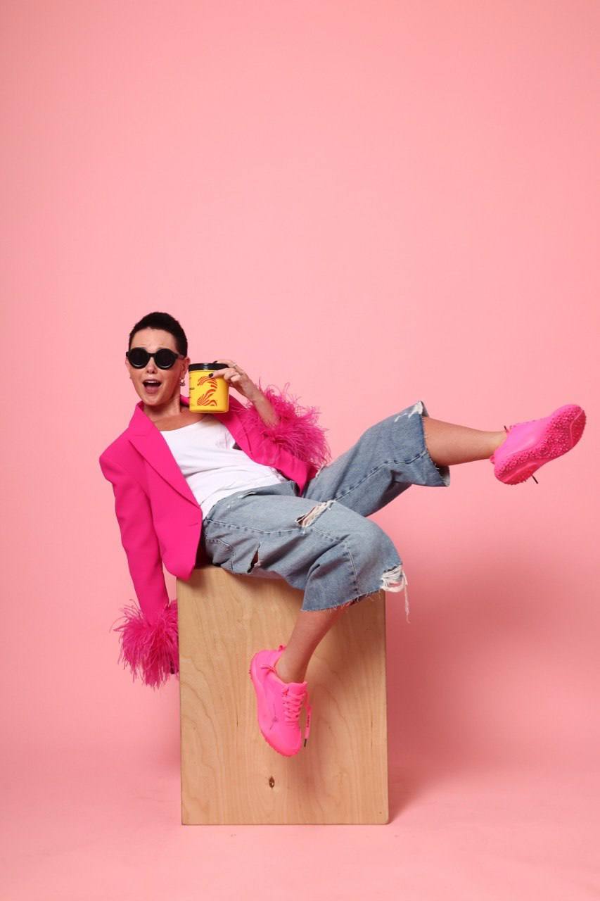 Person sitting on a wooden block with pink background, wearing pink jacket, sunglasses, gray pants, and pink shoes, smiling.